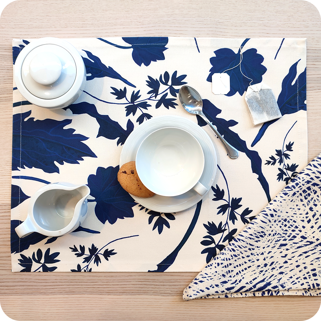 Tovaglietta rettangolare apparecchiata realizzata con un tessuto stampato con una fantasia di foglie blu su fondo chiaro; tovagliolo in lino abbinato con un motivo a tratteggi bianchi su fondo blu