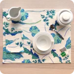 Tovaglietta rettangolare apparecchiata realizzata con un tessuto stampato con una fantasia con elementi foglie verdi azzurre su fondo chiaro; tovagliolo in lino abbinato