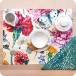Tovaglietta rettangolare apparecchiata realizzata con un tessuto stampato con una fantasia fiori, coccinelle e farfalle; tovagliolo abbinato in lino con un motivo a tratteggi verdi su fondo chiaro