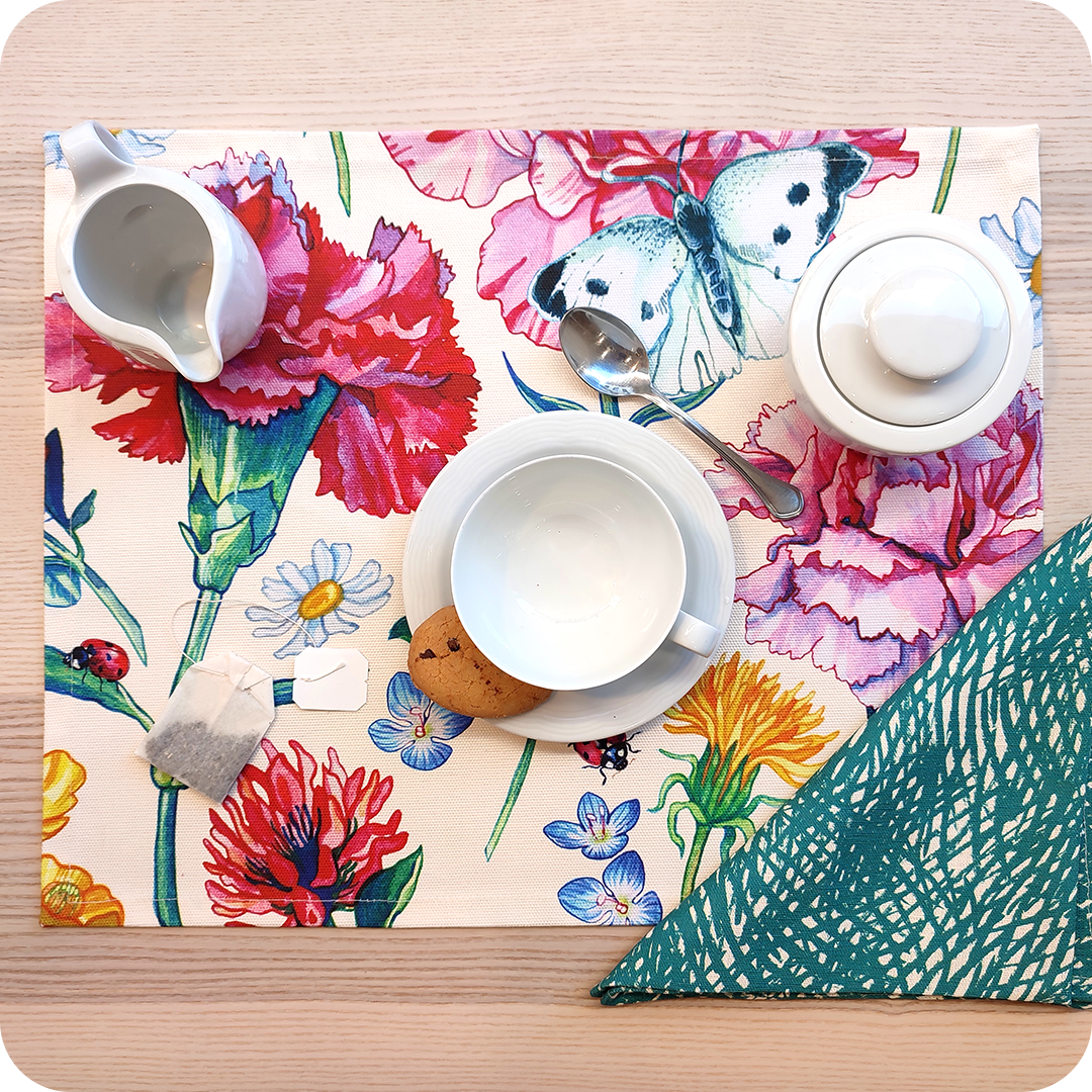 Tovaglietta rettangolare apparecchiata realizzata con un tessuto stampato con una fantasia fiori, coccinelle e farfalle; tovagliolo abbinato in lino con un motivo a tratteggi verdi su fondo chiaro