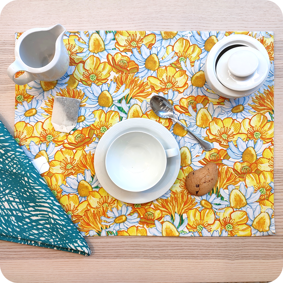 Tovaglietta rettangolare apparecchiata realizzata con un tessuto stampato con una fantasia fiori bianchi e gialli; tovagliolo in lino abbinato stampato con un motivo a tratteggi verdi su fondo chiaro