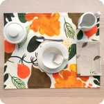 Tovaglietta rettangolare apparecchiata realizzata con un tessuto stampato con una fantasia con elementi vegetali stilizzati con colori caldi su fondo chiaro; tovagliolo abbinato in lino écru stampato con pigne e rametti di pino