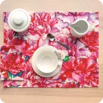 Tovaglietta rettangolare apparecchiata realizzata con un tessuto stampato con una fantasia con garofani rosa e magenta; tovagliolo in lino abbinato