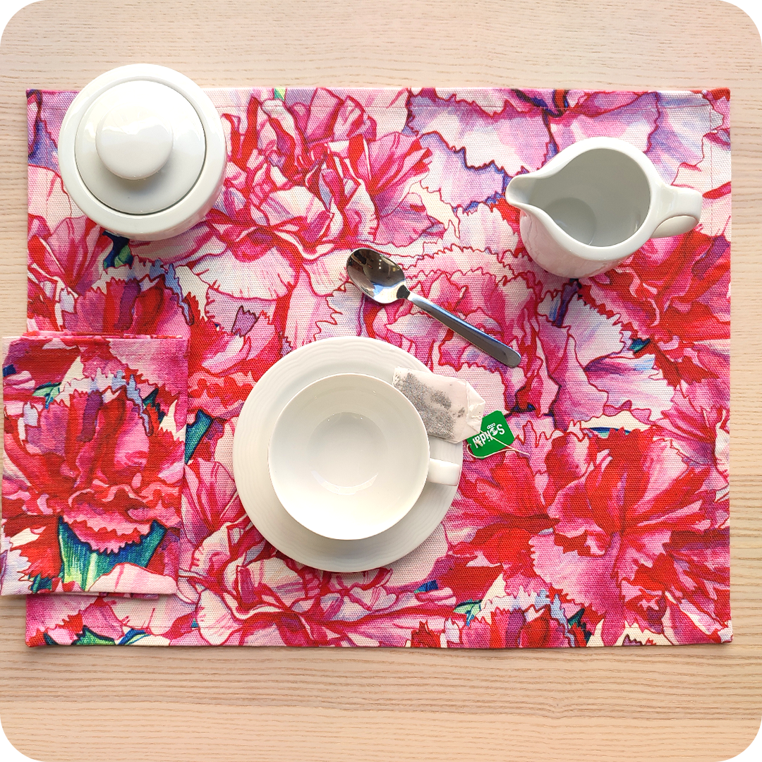 Tovaglietta rettangolare apparecchiata realizzata con un tessuto stampato con una fantasia con garofani rosa e magenta; tovagliolo in lino abbinato