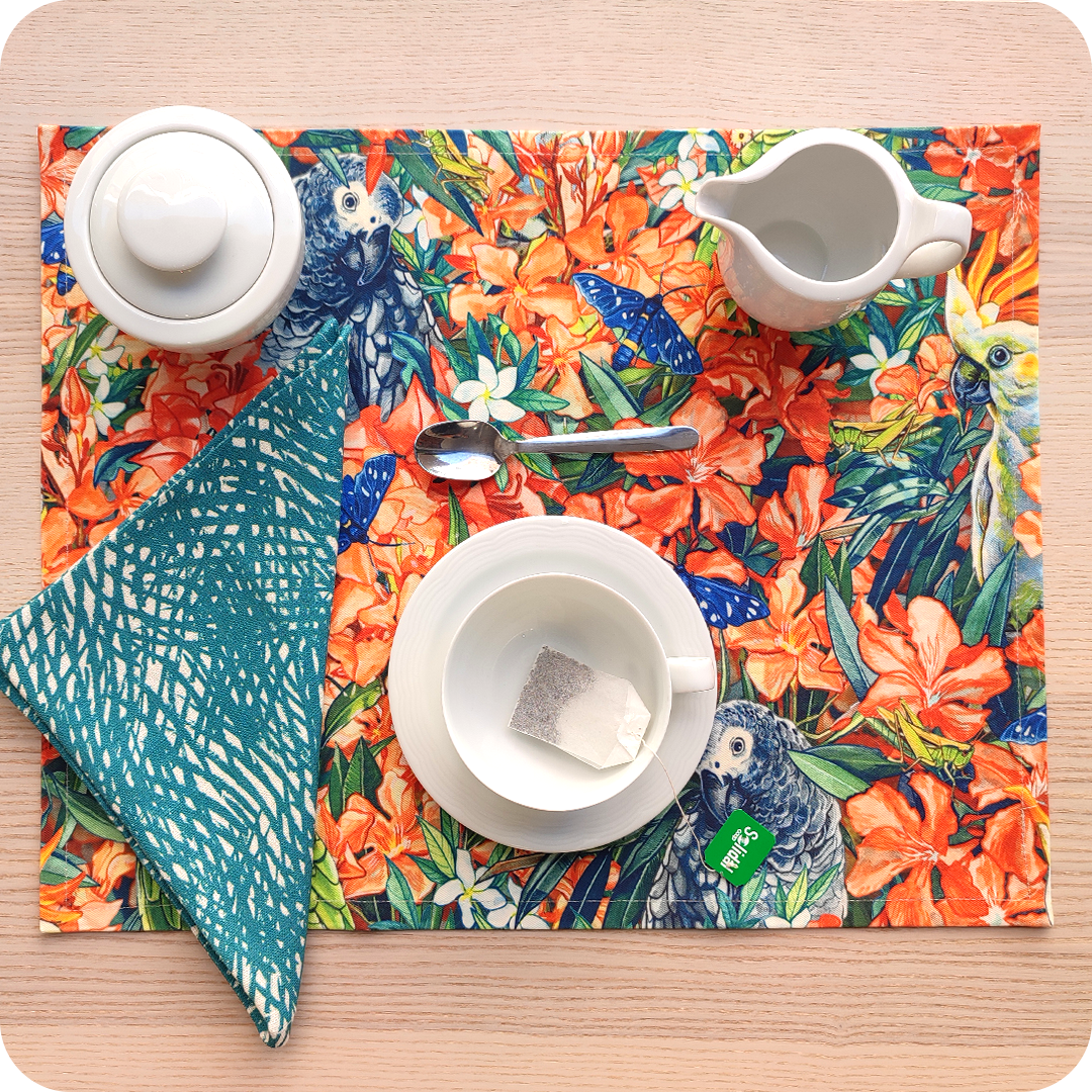 Tovaglietta rettangolare apparecchiata realizzata con un tessuto stampato con una fantasia con pappagalli, falene, grilli che sbucano da un cespuglio di fiori; tovagliolo in lino abbinato con motivo a tratteggio verde e bianco