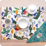 Tovaglietta rettangolare apparecchiata realizzata con un tessuto stampato con una fantasia con lucertole, fiori e foglie su fondo chiaro; tovagliolo in lino abbinato on motivo a tratteggio verde su fondo chiaro
