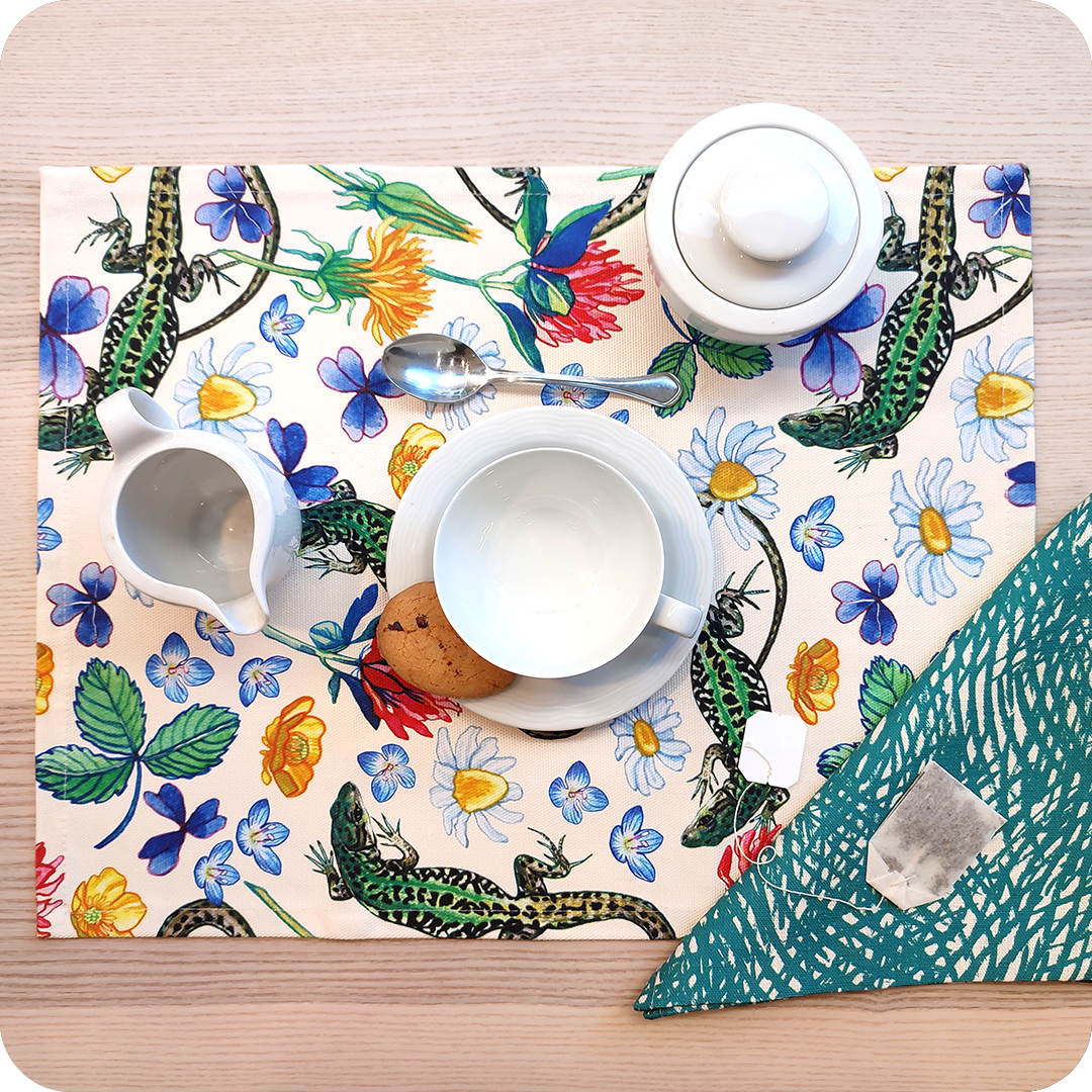 Tovaglietta rettangolare apparecchiata realizzata con un tessuto stampato con una fantasia con lucertole, fiori e foglie su fondo chiaro; tovagliolo in lino abbinato on motivo a tratteggio verde su fondo chiaro