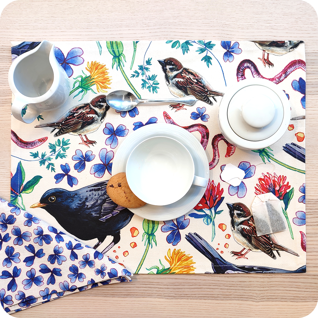 Tovaglietta rettangolare apparecchiata realizzata con un tessuto stampato con una fantasia con merli, passerotti, lombrichi, fiori, foglie su fondo chiaro; tovagliolo abbinato in lino con trifogli blu su fondo chiaro