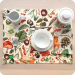 Tovaglietta rettangolare apparecchiata realizzata con un tessuto stampato con una fantasia con disegni di pettirossi, funghi, foglie, pigne, ghiande, fiori e bacche; tovagliolo in lino abbinato