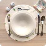 Tovaglietta tonda apparecchiata realizzata in tessuto stampato con una fantasia con rametti di pino e pigne; tovagliolo abbinato in lino naturale