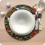 Tovaglietta tonda apparecchiata realizzata in tessuto stampato con una fantasia con bacche e foglie; tovagliolo abbinato in lino écru con pattern di rametti di pino e pigne
