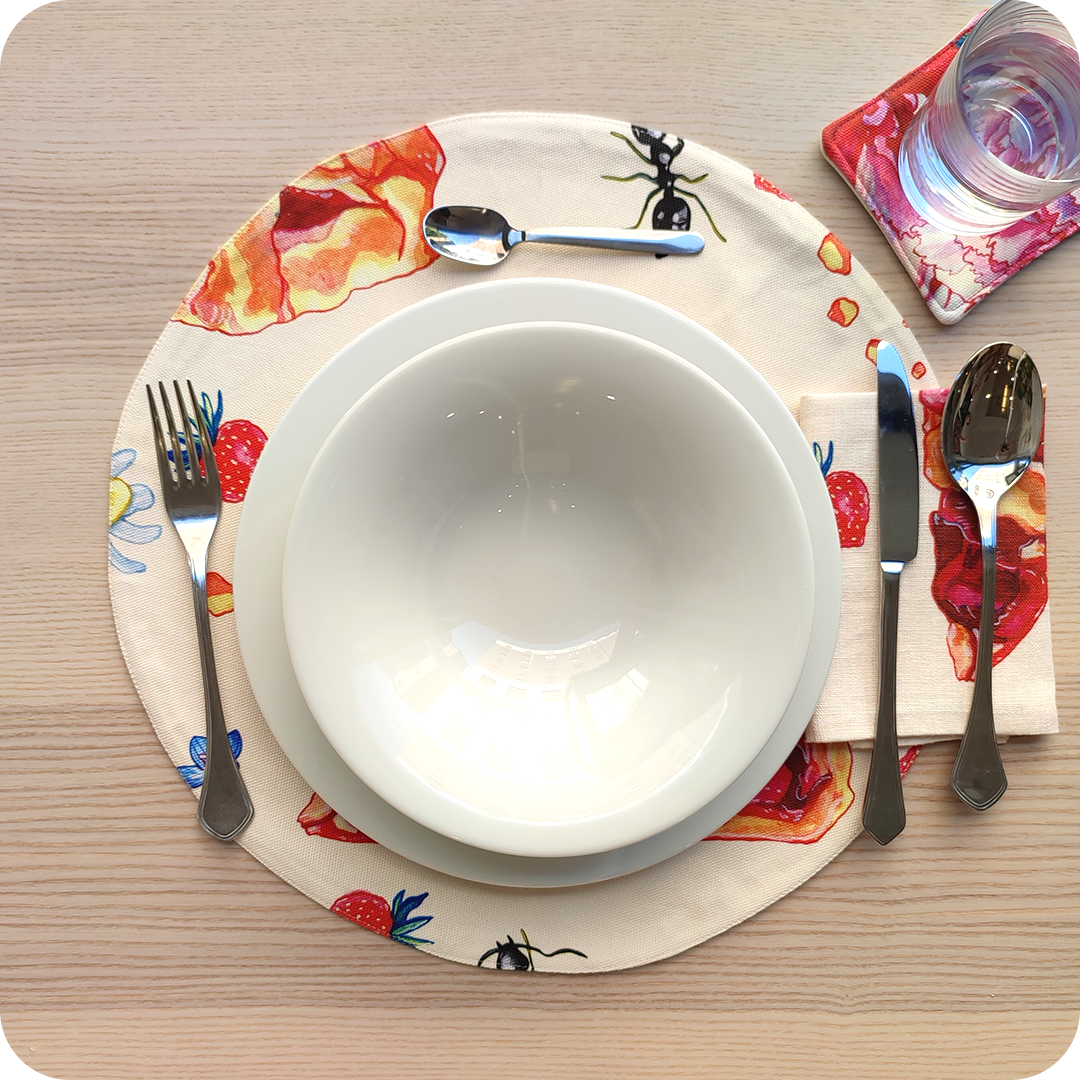 Tovaglietta tonda apparecchiata realizzata in tessuto stampato con una fantasia con fette di crostata, fiori, formiche e fragole su fondo chiaro; tovagliolo in lino abbinato