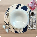 Tovaglietta tonda apparecchiata realizzata in tessuto stampato con fantasia di foglie blu su fondo chiaro; tovagliolo in lino abbinato con tratteggi bianchi su fondo blu