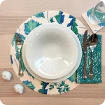 Tovaglietta tonda realizzata in tessuto stampato con fantasia di foglie verdi e azzurre su fondo chiaro; tovagliolo abbinato in lino con motivo a tratteggio verde su fondo chiaro