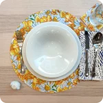 Tovaglietta tonda apparecchiati realizzata in tessuto stampato con fantasia fiori bianchi e gialli; tovagliolo in lino abbinato con tratteggi bianchi su fondo blu