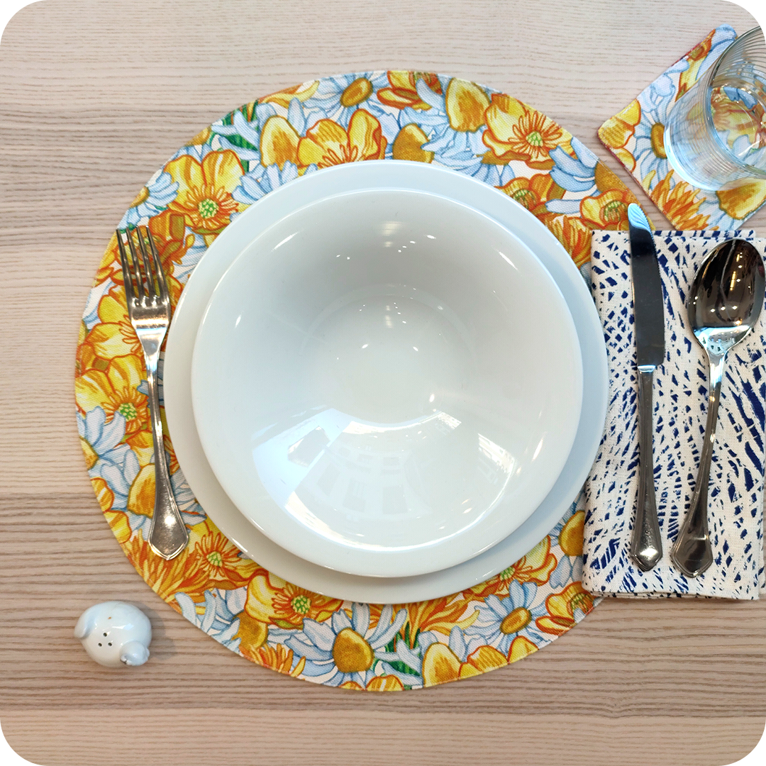 Tovaglietta tonda apparecchiati realizzata in tessuto stampato con fantasia fiori bianchi e gialli; tovagliolo in lino abbinato con tratteggi bianchi su fondo blu