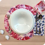 Tovaglietta tonda apparecchiata realizzata in tessuto stampato con fantasia con garofani sui toni del rosa e del magenta; tovagliolo in lino abbinato con trifogli blu su fondo bianco