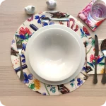 Tovaglietta tonda apparecchiata realizzata in tessuto stampato con una fantasia con merli, passerotti, lombrichi, fiori e foglie su fondo chiaro; tovagliolo in lino abbinato