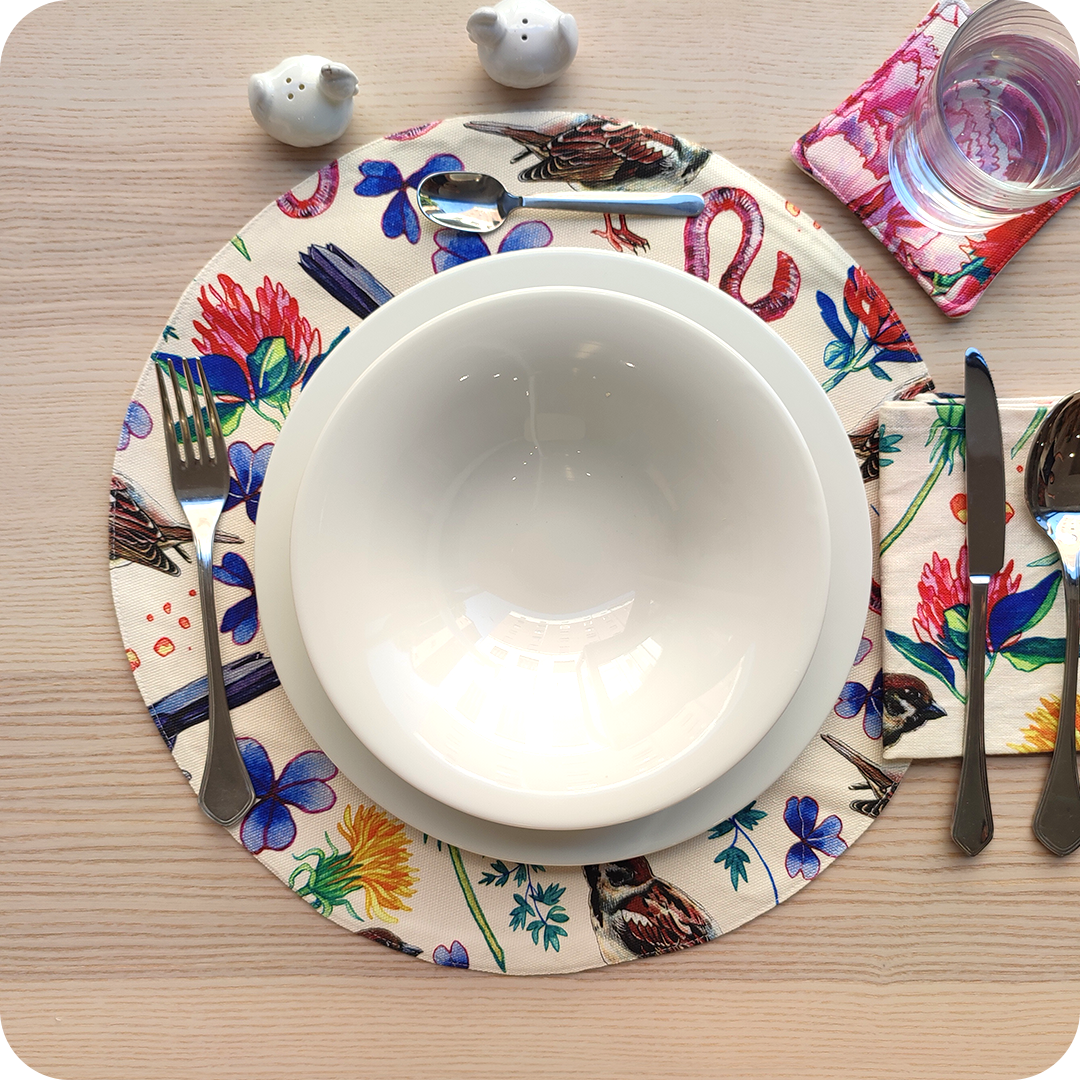 Tovaglietta tonda apparecchiata realizzata in tessuto stampato con una fantasia con merli, passerotti, lombrichi, fiori e foglie su fondo chiaro; tovagliolo in lino abbinato
