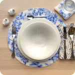 Tovaglietta tonda apparecchiata realizzata in tessuto stampato con una fantasia con grappoli d'uva, rose canine e farfalle sui toni dell'azzurro; tovagliolo abbinato in lino stampato con motivo a tratteggio bianco e blu