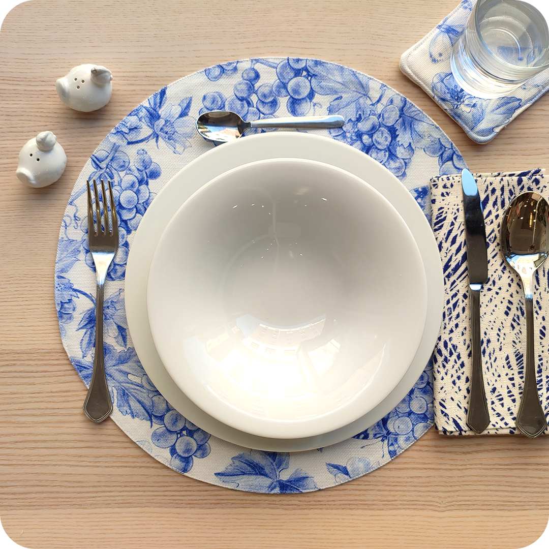 Tovaglietta tonda apparecchiata realizzata in tessuto stampato con una fantasia con grappoli d'uva, rose canine e farfalle sui toni dell'azzurro; tovagliolo abbinato in lino stampato con motivo a tratteggio bianco e blu