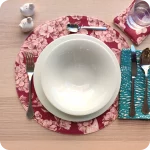 Tovaglietta tonda apparecchiata realizzata col fronte in tessuto stampato con una fantasia con grappoli d'uva, rose canine e farfalle sui toni del bordeaux; tovagliolo abbinato in lino con motivo a tratteggio verde e bianco