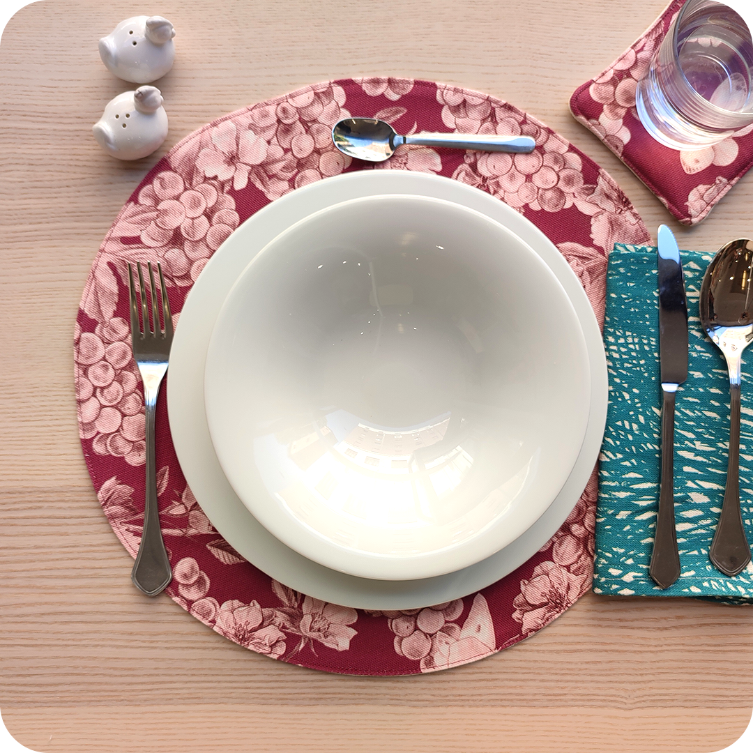 Tovaglietta tonda apparecchiata realizzata col fronte in tessuto stampato con una fantasia con grappoli d'uva, rose canine e farfalle sui toni del bordeaux; tovagliolo abbinato in lino con motivo a tratteggio verde e bianco