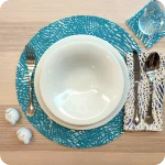 Tovaglietta tonda realizzata in tessuto stampato con un motivo a tratteggio azzurro su fondo chiaro; tovagliolo in lino abbinato con tratteggi bianchi su fondo blu