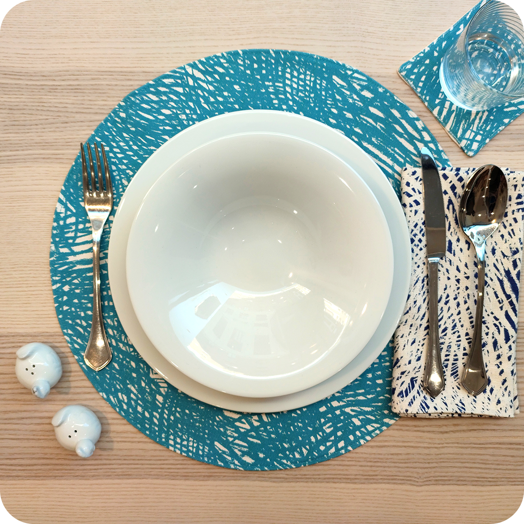 Tovaglietta tonda realizzata in tessuto stampato con un motivo a tratteggio azzurro su fondo chiaro; tovagliolo in lino abbinato con tratteggi bianchi su fondo blu