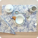 Tovaglietta rettangolare apparecchiata realizzata con un tessuto stampato con una motivo di tratteggi bianchi su fondo blu; tovagliolo in lino abbinato