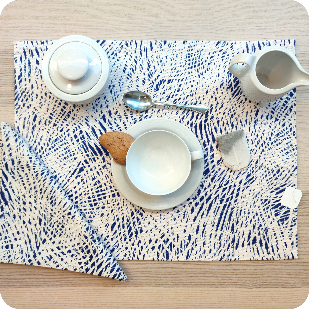 Tovaglietta rettangolare apparecchiata realizzata con un tessuto stampato con una motivo di tratteggi bianchi su fondo blu; tovagliolo in lino abbinato
