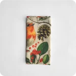 Tovagliolo quadrato piegato in lino écru stampato con una fantasia di pettirossi, pigne, rametti, fiori, funghi, ghiande, bacche, foglie