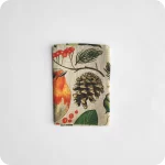 Tovagliolo quadrato piegato in lino écru stampato con una fantasia di pettirossi, pigne, rametti, fiori, funghi, ghiande, bacche, foglie